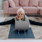 A senior woman enjoys a relaxing yoga session indoors, stretching while using her laptop.
