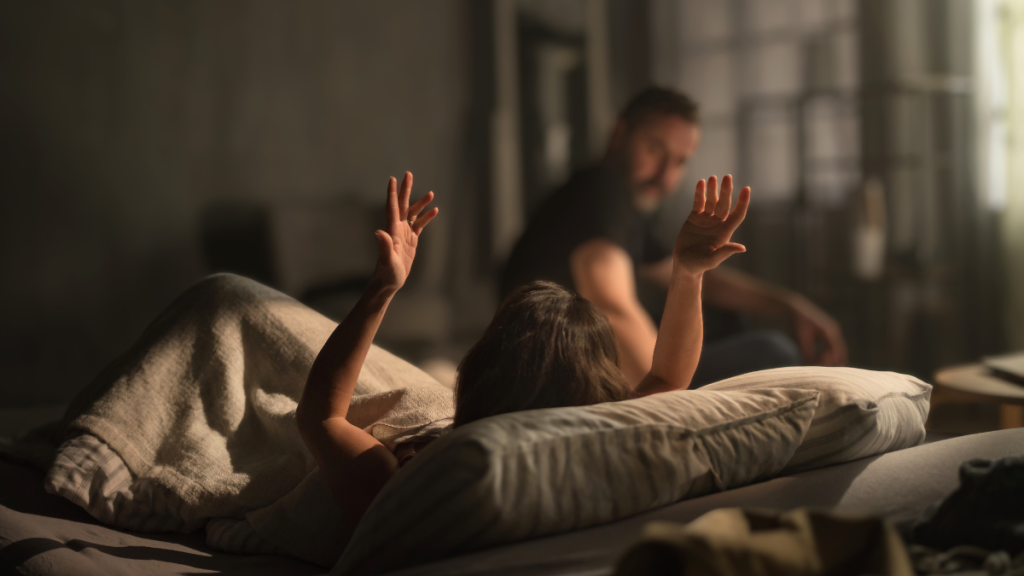 Een stel in een slaapkamer. De vrouw houdt gefrustreerd haar handen omhoog